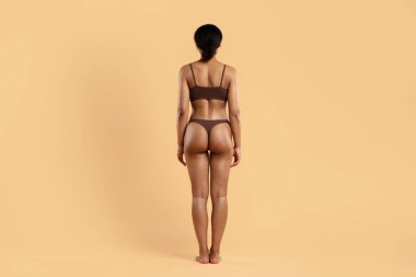 Kahverengi iç çamaşırlı, bej arka planda genç siyahi bir kadın. Formda kıvrımlar, sağlam bacaklar ve tanımlanmış bel fitness hedefleri, vücut bakımı rutinleri ve kilo verme ilerlemesini vurguluyor