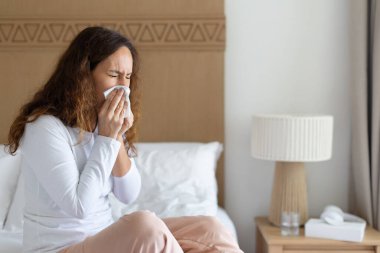 Yetişkin bir kadın yatakta yatarken mendile hapşırıyor. Soğuk ya da alerji kavramı, yaşam tarzı sağlık sorunu, evde bakım, hastalık önleme ve kapalı alanda refah anı