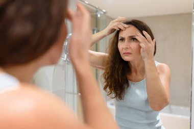 Yetişkin bir kadın banyo aynasına bakarken alnındaki çizgileri parmaklarıyla kontrol ediyor. Yaşlanan cilt analizi, rutin cilt bakımı ve evde gerçekçi güzellik bakımı.