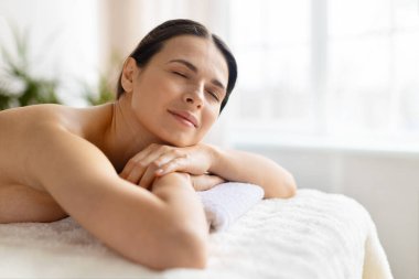 Gülümseyen kadın masaj masasında dinleniyor, spa tedavisinden sonra, kendini yenilenmiş ve sakin hissediyor. Işıl ışıl iç mekanlarda sağlık, güzellik, stres rahatlama ve sağlıklı yaşam tarzı kavramı