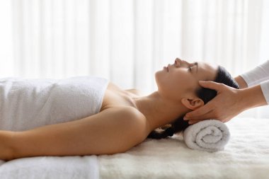 Genç, güzel bir kadın lüks bir kaplıcada kafa masajından zevk alıyor, gözleri kapalı bir tedavi yatağında uzanıyor, sakin bir atmosfer, sağlığa, cilt bakımına odaklanıyor,