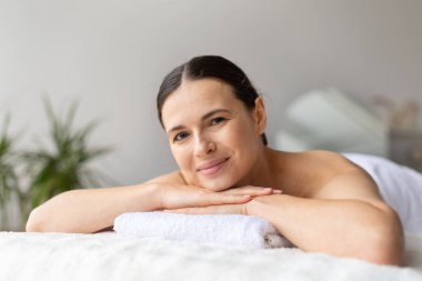 Gülümseyen güzel bir kadın spa masasında yatıyor, masaj tedavisini bekliyor, parlak minimalist iç mekan refahı, rahatlığı ve lüks kişisel bakım kavramını vurguluyor.