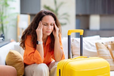 Yetişkin beyaz kadın, otel lobisinde bavulun yanında otururken hayal kırıklığı içinde başını tutuyor. Seyahat stresi ya da modern iç mekan yaşam tarzında baş ağrısı kavramı.