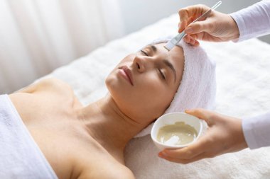 Beyaz kadın huzur içinde yatarken, yüz maskesi kaplıcada, gözleri kapalı, iç kısımları en az parlak cilt bakımını, sağlık terapisini ve rahatlamayı ön plana çıkarıyor.