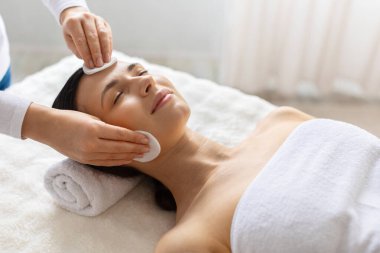 Kozmetik doktoru, yüz terapisi sırasında pamukla kaplıcaya uzanan genç bir kadının yüzünü temizliyor. Profesyonel eller cilt bakımı yapıyor.