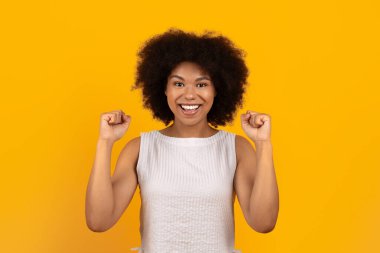 Kutlamak için yumruklarını kaldıran neşeli genç siyahi kadın, sarı arka planda stüdyo yaşam tarzı portresi başarıyı, heyecanı, mutluluğu, başarıyı ve olumlu duyguları ifade ediyor.