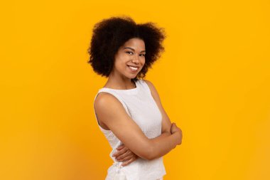 Stüdyoda kollarını kavuşturarak güvenle duran gülümseyen genç siyahi kadın, sarı arka plan, yaşam tarzı portresi pozitifliği, güveni, başarıyı ve modern gündelik tarzını ifade ediyor.