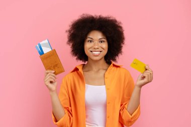 Gülümseyen genç siyah kadın elinde pasaport ve kredi kartı, seyahat ödemesi ve rezervasyon konsepti, pembe arka plan, turizm finans ve uluslararası yaşam tarzı teması ile izole edilmiş