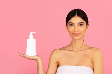 Genç bir kadın açık avuçla pembe arka planda kozmetik şişesini sunuyor. Cilt ürünleri modeli, güzellik reklamcılığı ve markalaşma kavramı