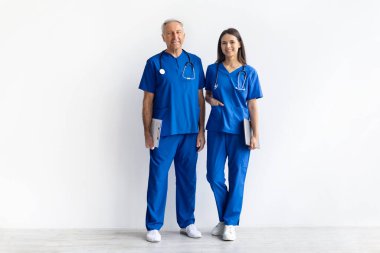Kıdemli erkek doktor ve genç bayan doktor birlikte mavi önlükle beyaz stüdyo geçmişi, hastane ve klinik hizmetleri için profesyonel sağlık ekibi portresi üzerinde duruyorlar.