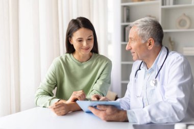 Üst düzey erkek doktor, modern klinikteki konsültasyon, dostane tartışma ve sağlık hizmetleri iletişim konsepti sırasında genç kadın hastaya tıbbi sonuçları açıklıyor.