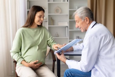 Hamile kadın, doğum öncesi konsültasyon, annelik planlaması ve sağlık belgelendirme konsepti sırasında kıdemli erkek doktorla birlikte tıbbi belgeleri inceliyor.