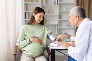 Hamile kadının doğum öncesi muayenesi, doğum öncesi sağlık kontrolü ve rutin hamilelik bakımı sırasında kan basıncını ölçen kıdemli erkek doktor.