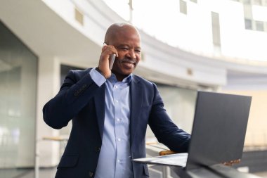 Ofis korkuluklarında dizüstü bilgisayar kullanırken akıllı telefondan konuşan, modern kurumsal alandaki dijital iş ve iş iletişimini birleştiren, gülümseyen zenci, olgun iş adamı.