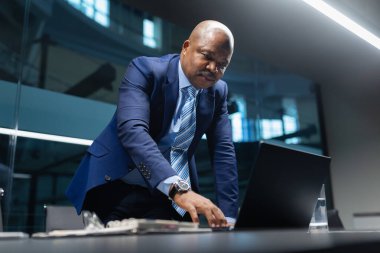 Bilgisayarla çalışırken konferans masasına yaslanan, modern bir şirket ofisinde kararlılık, liderlik ve pratik kararlar veren, siyahi, olgun iş adamı.