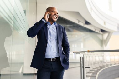 Ofis koridorunda dikilirken akıllı telefondan konuşan olgun, gülümseyen zenci işadamı, modern, parlak bir şirket ortamında gevşemiş yönetici iletişimi.