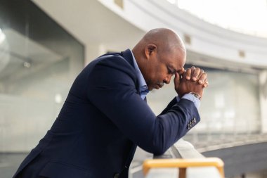 Stresli, olgun siyahi işadamı, modern ofiste iş günü talep ederken parmaklıklara yaslanıyor, baskı ve derin düşünceleri yansıtıyor.