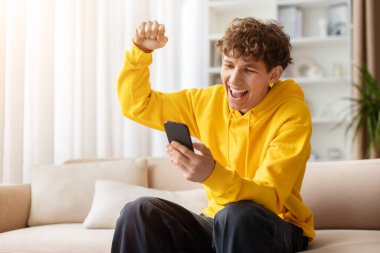 Heyecanlı genç adam evde akıllı telefona bakarken başarıyı kutluyor, yumruğunu kaldırıyor, sarı kapüşonlu giyiyor, zaferini, başarısını ve içeride olumlu duygularını ifade ediyor.