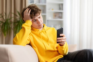 Sarı kapüşonlu düşünceli genç adam evde akıllı telefonu kontrol ediyor. Stresli ya da endişeli ifade, dijital iletişim ve modern yaşam tarzı problemi kavramı.