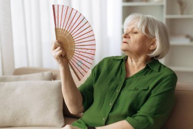 Yaşlı, beyaz bir kadın evdeki kanepede dinlenirken bir vantilatörle kendini serinletiyor. Olgun bir kadın yaz boyunca sıcaktan rahatsız oluyor.