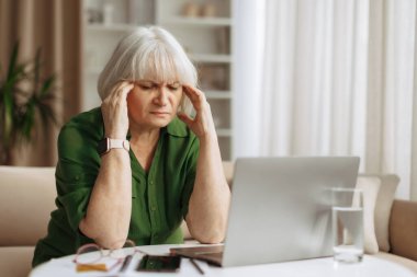 Evde dizüstü bilgisayar başında çalışırken başını kapatan yaşlı bir kadın. Stres ya da baş ağrısı çeken yaşlı bir kadın. Günlük emeklilik yaşam tarzında zorlu bir dönem.
