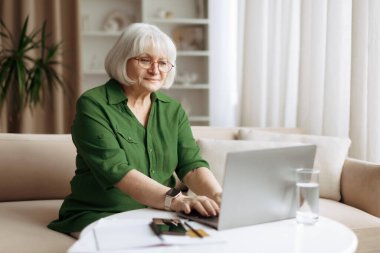 Evde dizüstü bilgisayarda çalışan yaşlı beyaz kadın, dijital teknoloji, emeklilik aktivitesi, uzaktan iletişim ve günlük konfor ile sakin bir son sınıf yaşam tarzı sahnesi