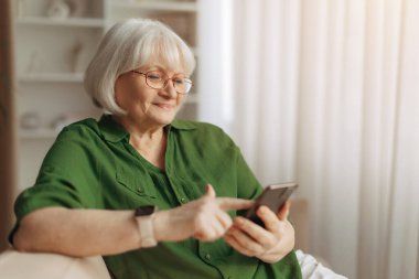 Evde koltukta otururken akıllı telefon kullanan yaşlı beyaz kadın, son sınıf öğrencisi kadın cep telefonu teknolojisi ve modern emeklilik yaşam tarzıyla güvenli bir şekilde etkileşimde bulunuyor.