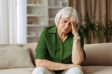 Yaşlı, beyaz bir kadın, evinde koltukta otururken başını tutuyor. Yaşlı bir kadın yorgunluk ya da baş ağrısı çekiyor. Huzurlu bir emeklilik hayatı ve duygusal refah kavramı..