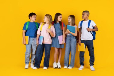 Sarı stüdyo arka planında izole edilmiş not defterlerini tutarken, genç öğrenciler birbirleriyle etkileşime geçip sohbet ediyorlar. Arkadaşlığı, iletişimi, okul hayatını ve eğitimi gösteren doğal duygular