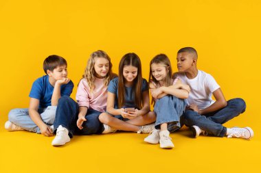 Bir grup çocuk birlikte oturuyor ve sarı arka planda akıllı telefon kullanıyorlar. Dijital çocukluk, teknoloji, iletişim ve modern eğitim kavramı