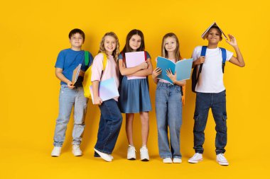 Sarı stüdyo arka planında not defterleri ve sırt çantalarıyla poz veren bir grup çocuk. Neşeli ifadeler öğrenme, yaratıcılık, eğitim ve pozitif okul atmosferini yansıtıyor
