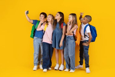 Küçük çocuklar sarı stüdyo arka planında selfie çekiyorlar, gülümsüyorlar ve eğleniyorlar. Modern okul yaşam tarzı, arkadaşlık, teknoloji ve eğitim kavramı