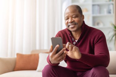 Evde kanepede otururken akıllı telefon kullanarak gülümseyen olgun bir zenci. İletişim, teknoloji ve modern yaşam ortamlarını gösteren sıradan bir ev hayatı.