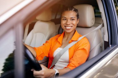 Genç siyahi kadın araba kullanıyor ve kameraya güvenle gülümsüyor, emniyet kemeri takıyor ve şehir içi yolculuğun keyfini çıkarıyor.