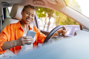 Gülümseyen genç siyah kadın araba kullanıyor ve elinde akıllı telefona bakıyor, modern bağlantılı yaşam tarzını ve dikkati dağılmış riskli sürüş konseptini gösteriyor.