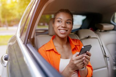 Arka koltukta rahat bir şekilde uzanan genç siyahi kadın akıllı telefon tutuyor ve gülümsüyor, güneşli gündüz araba yolculuğu sırasında rahatlıyor.