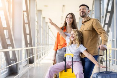 Havaalanı koridorunda bavulun üzerine oturmuş gülümseyen anne babasıyla aile tatili yolculuğunun keyfini çıkarırken kollarını açan oyuncu kız.