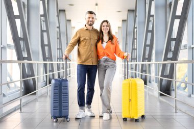 Genç çift havaalanında valizleriyle yürüyor, gülümsüyor ve tatil seyahatlerine hazırlanıyor, ayrılmadan önce modern terminal atmosferinin tadını çıkarıyorlar.