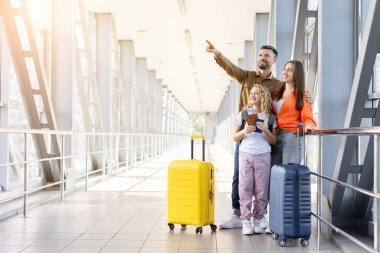 Havalimanı koridorunda renkli bavulları olan gülümseyen aile baba, tatil seyahatine ve birlikte heyecanlı bir tatil yolculuğuna hazırlanırken ileri doğru işaret ediyor.