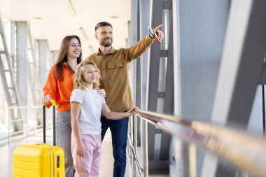 Havalimanı penceresinden bakan aile, elinde pasaport tutarken pisti gösteren baba, tatil ve seyahat macerasına hazır.