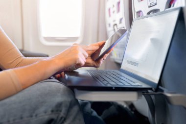 Yetişkin beyaz kadının uçuş sırasında dizüstü bilgisayar tepsisi üzerinde akıllı telefon kullanmasına yakın çekim. Uçak kabininde iş seyahati ve ulaşım kavramı