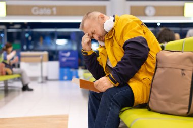 Havalimanı salonunda elinde pasaportla oturan yorgun, beyaz bir erkek yolcu. Bagaj ve kulaklıkla seyahat gecikmesi ve ulaşım bekleme kavramı