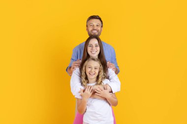 Üç kişilik mutlu aile, parlak sarı stüdyo arka planında birlikte poz veriyor, kameraya gülümsüyor ve kucaklaşıyorlar. Neşeli ebeveynler ve çocuklar sıcak yaşam tarzının tadını çıkarıyorlar.