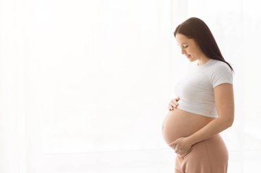 Aydınlık bir evde karnını tutan hamile bir kadının yan görüntüsü. Sağlıklı hamilelik, doğum öncesi yaşam tarzı ve bebek bekleyen barışçıl annelik kavramı.