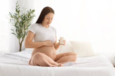 Hamile bir kadın yatakta bacak bacak üstüne atmış, elinde sütle evde karnına dokunuyor. Doğum öncesi beslenme, sağlıklı hamilelik yaşam tarzı ve annelik refahı kavramı
