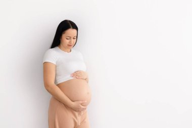 Hamile güzel bir kadın beyaz duvar arkasında duruyor, karnını nazikçe tutuyor, anne bakımını ve sağlıklı hamileliği sembolize ediyor, kopyalama alanını.