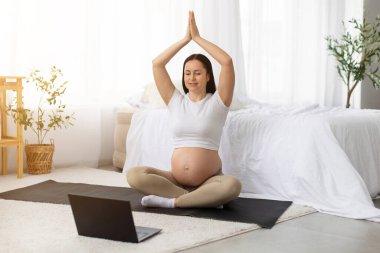 Genç hamile bir kadın doğum öncesi yoga seansında elleri havaya kaldırıyor. Yanında dizüstü bilgisayarla. Çevrimiçi fitness eğitimi ve sağlıklı annelik yaşam tarzı konsepti