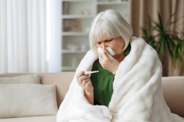 Üst düzey kadın battaniyeye sarılıyken vücut ısısını termometreyle ölçüyor. Olgun beyaz kadın soğuk algınlığı ya da evde grip belirtileri gösteriyor.