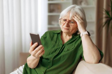 Gözlüklü yaşlı kadın evde otururken endişeli bir ifadeyle akıllı telefona bakıyor. Olgun Kafkas kadın okuma iletisi ya da mobil aygıt üzerinde haberler