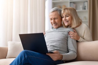 Üst düzey bir çift parlak bir oturma odasında dizüstü bilgisayar kullanarak kanepede dinleniyor. Olgun erkek ve kadın çevrimiçi aktivitelerden keyif alarak modern dijital yaşam tarzını ve evdeki aile rahatlığını temsil ediyorlar.